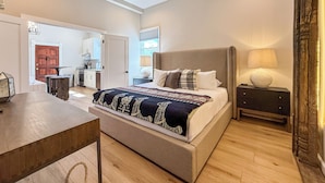 A spacious bedroom with a large bed, a ceiling fan, a bedside table with a lamp, and a view of the kitchen and living area.