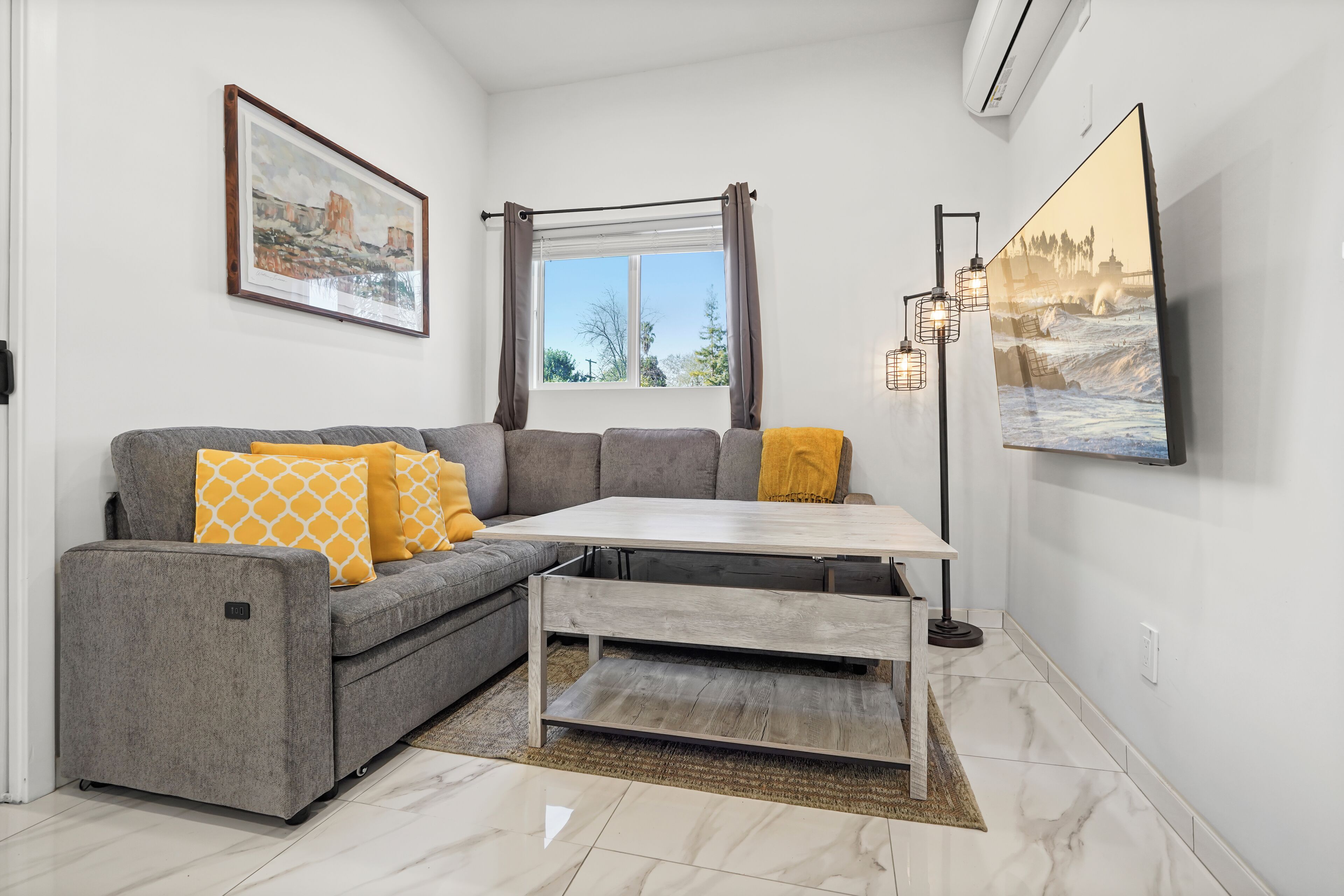 Un salón moderno con un sofá gris, cojines amarillos, una mesa de centro de madera, un ventanal con cortinas y un cuadro enmarcado en la pared.