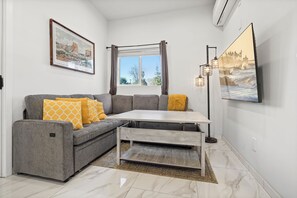 Ein modernes Wohnzimmer mit grauem Sofa, gelben Kissen, einem Holztisch, einem großen Fenster mit Vorhängen und einem gerahmten Gemälde an der Wand.