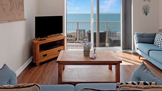Ein Wohnzimmer mit Couch, einem Couchtisch aus Holz, einem Fernsehgerät auf einem Holzboden und Blick auf das Meer durch eine Glastür.