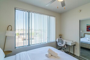 A hotel room with a bed, a desk, a chair, a lamp, and a large window with blinds. - Access 3104 (Tempe)