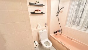 Ein kleines Badezimmer mit Toilette, Badewanne und Dusche. Über der Toilette befinden sich Regale und ein Fenster mit Vorhängen.