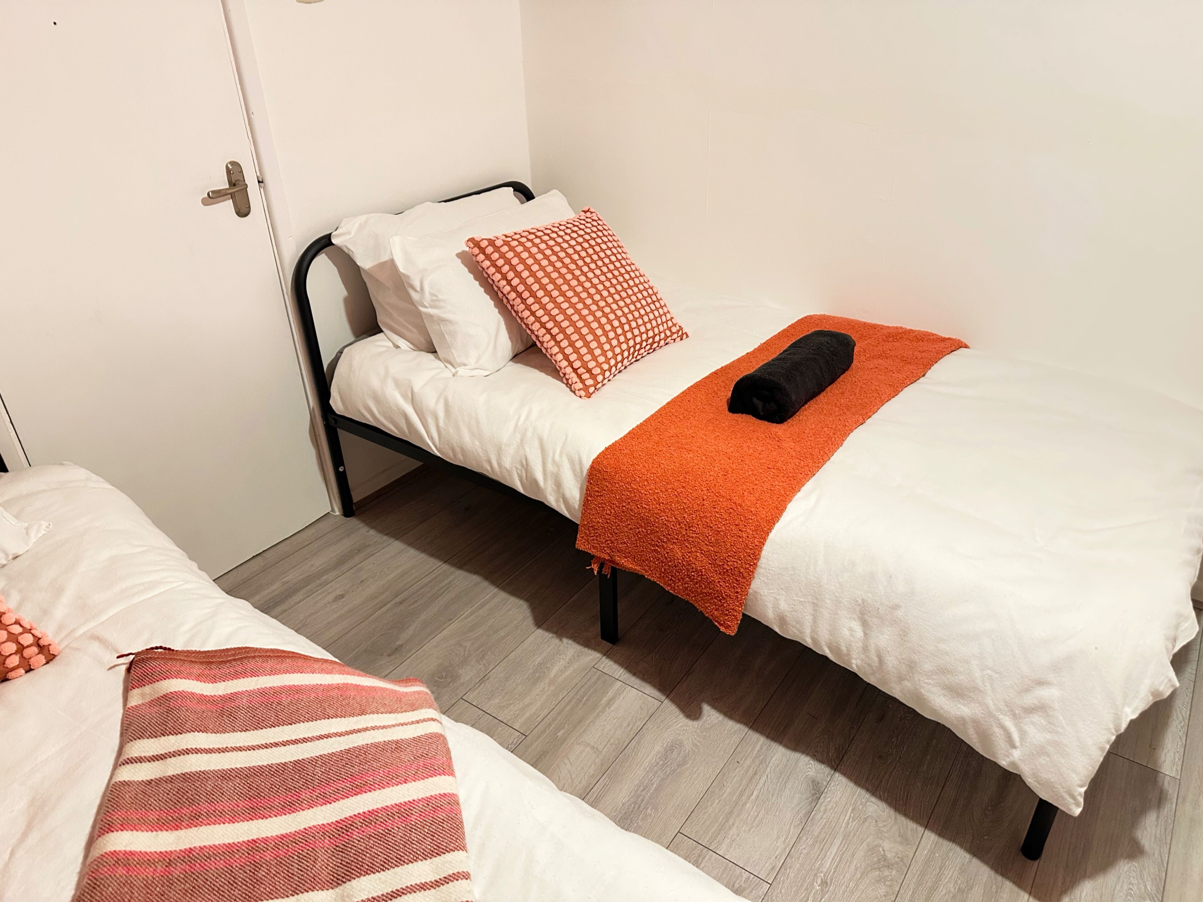 A single bed with white bedding, an orange blanket, and a black towel.