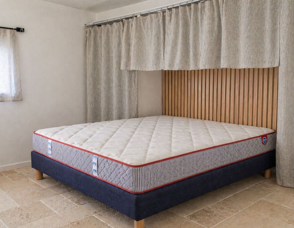 Een enkel bed met een gewatteerde matras, een houten hoofdbord en een gordijn.