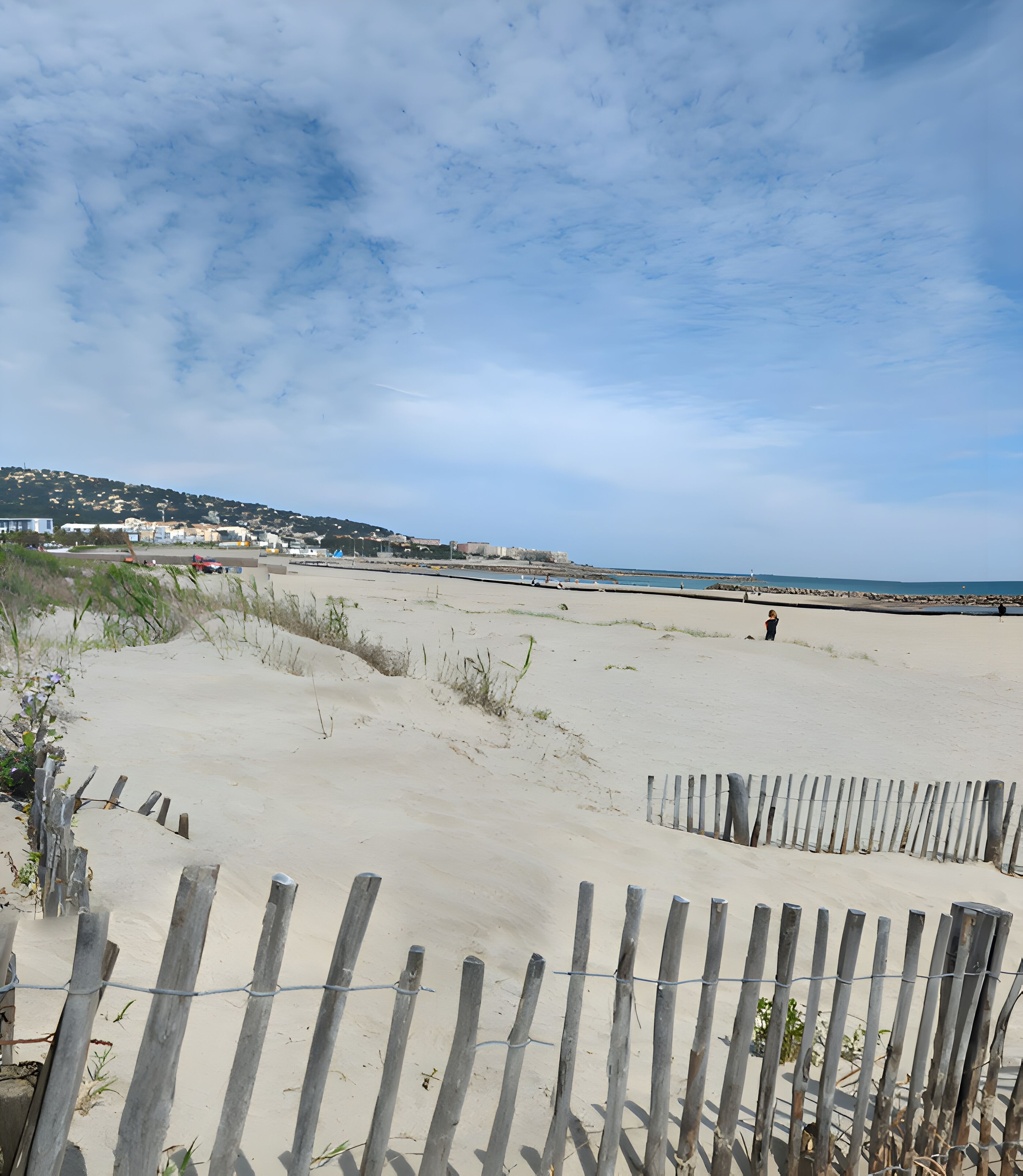 Une plage de sable bordée d’une clôture en bois, un ciel dégagé et des bâtiments qui se dessinent à l’horizon.