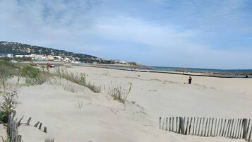 Une plage de sable bordée d’une clôture en bois, un ciel dégagé et des bâtiments qui se dessinent à l’horizon.