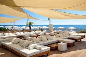 Un espace de détente aménagé au bord de la plage, offrant de nombreuses options d’assises, notamment des canapés et des fauteuils, le tout abrité sous de grands auvents.
