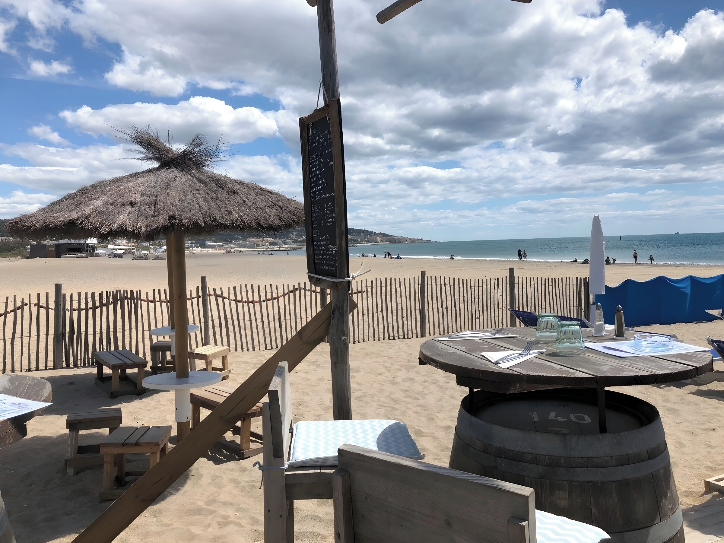 A beachside restaurant with outdoor seating, a thatched roof, and a view of the beach and ocean.