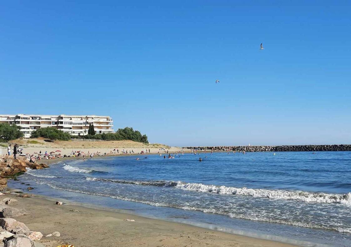 Une plage bordée d’une côte rocheuse, avec une jetée et un bâtiment doté de nombreux balcons.