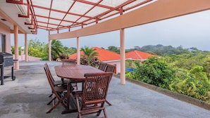 Une terrasse extérieure couverte, avec une table et des chaises en bois, un barbecue et une vue sur un paysage luxuriant, parsemé de maisons aux toits rouges.