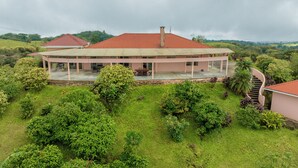 Une grande maison avec un toit rouge, entourée d’une végétation luxuriante et offrant une vue sur des collines ondulantes.