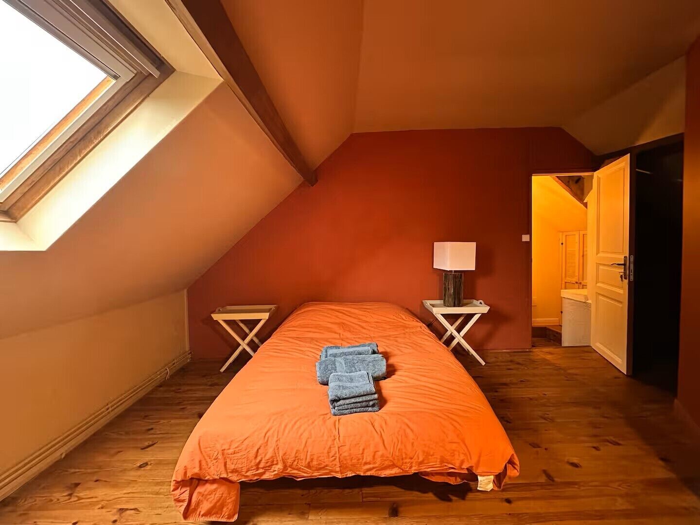Ein Schlafzimmer mit geneigter Decke, einem Bett mit orangefarbener Bettdecke, zwei Nachttischen mit Lampen und einer Tür zu einem anderen Raum.