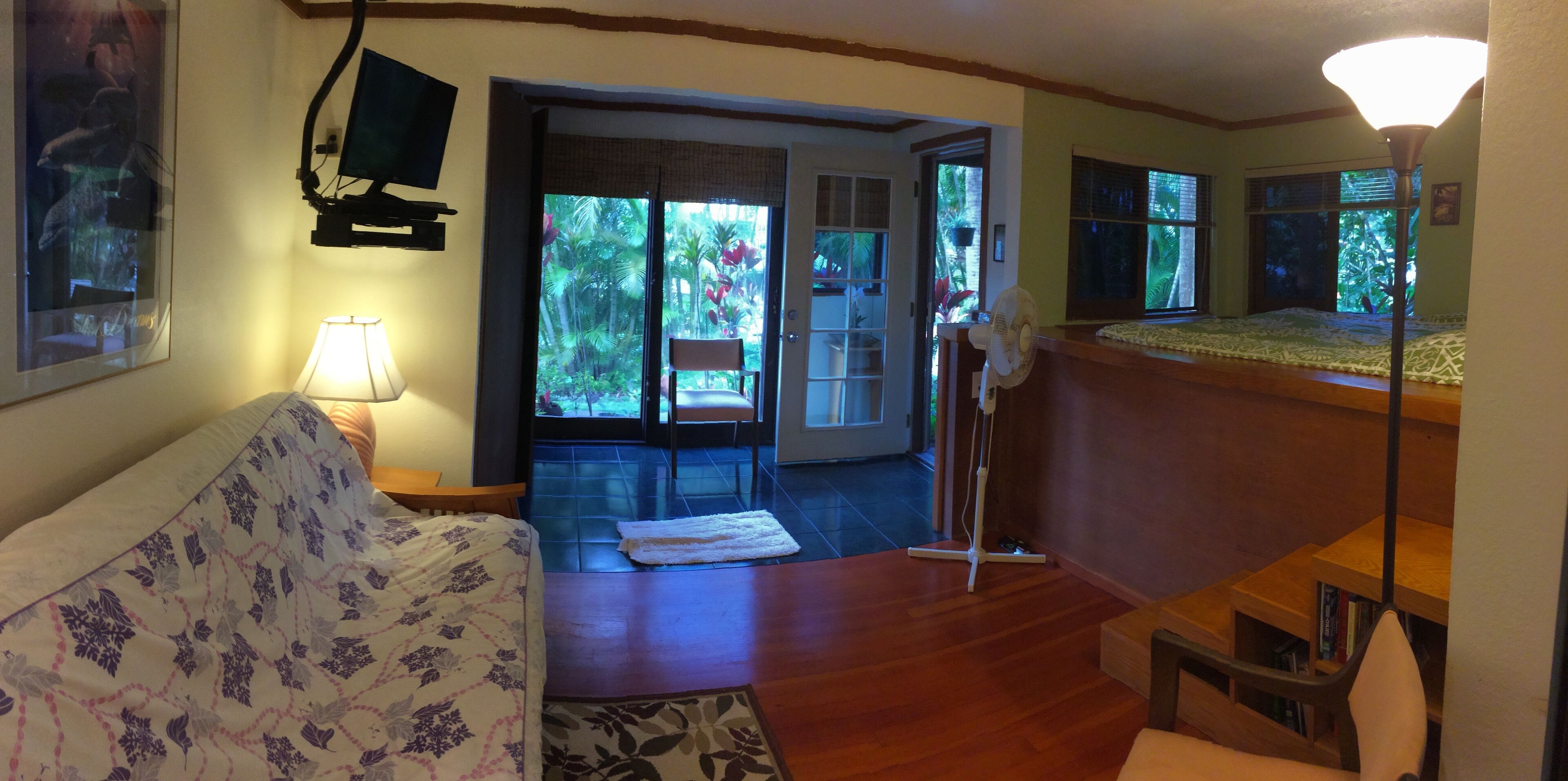 A room with a bed, a desk, a chair, a lamp, and a view of a balcony with plants.