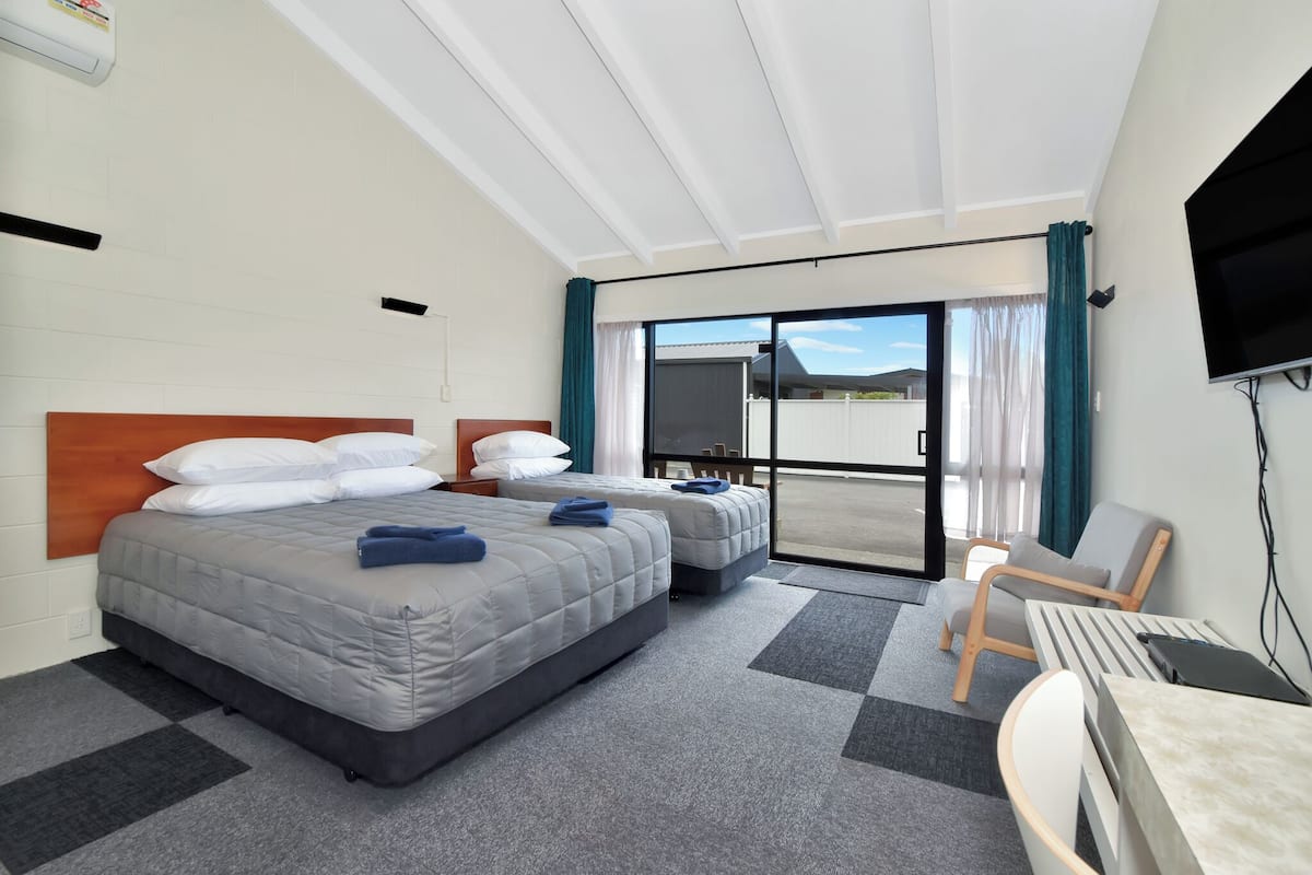 A hotel room with two beds, a desk, a chair, a TV, and a sliding glass door to the outside.