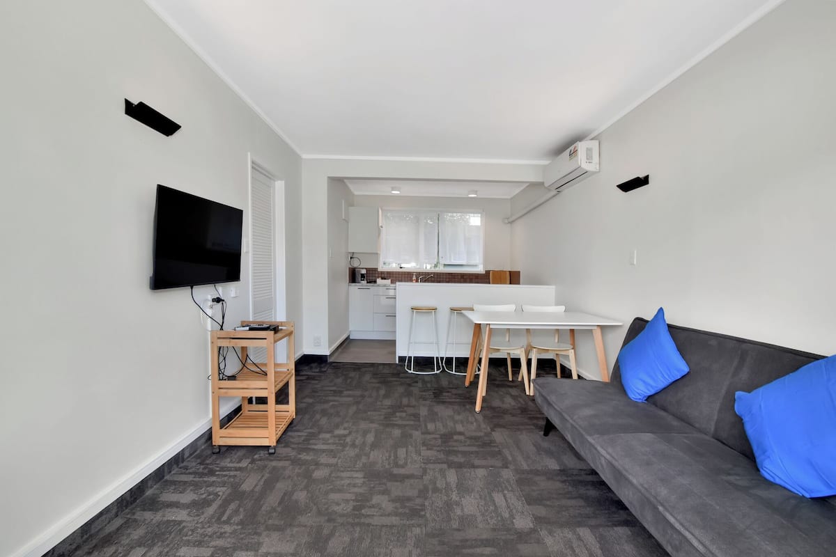 A modern living room with a grey sofa, a TV on the wall, a wooden TV stand, and a small dining area with a white table and stools.