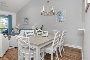 A dining area with a wooden table, white chairs, a chandelier, and framed artwork on the walls. - 2 BR Barefoot Resort Getaway - Free Shuttle to Beach - 2 Pools  (North Myrtle Beach)