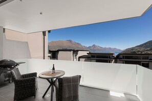 Ein Balkon mit Blick auf Berge und ein Gewässer. Dort steht ein Tisch mit einer Flasche und einem Glas, sowie zwei Korbstühle.