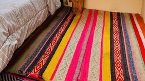 A cozy room with a bed, colorful striped carpet, a hanging decoration, and a view of greenery outside. - Salakantay glamp (Santa Teresa)