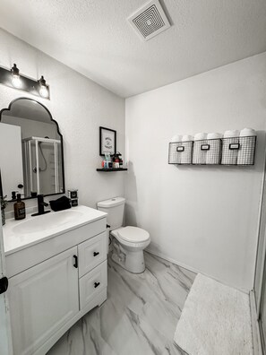 A bathroom with a white vanity, a toilet, a shower enclosure, and storage shelves.