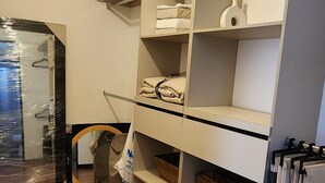 Una habitación con suelo de baldosas, un estante blanco con toallas dobladas y una cesta, y un bolso colgado.