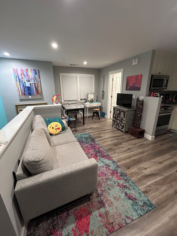 A living room with a grey sectional sofa, a colorful rug, a dining area with a table and chairs, a kitchen with a microwave and stove, and a TV on a cabinet.