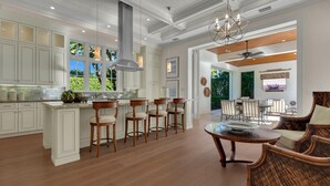 A spacious kitchen with white cabinetry, a central island with bar stools, and a dining area with wicker furniture. - Elevated Coastal Living | Luxury 4BR Home + Den | Designed for the Perfect Getaway (Naples)
