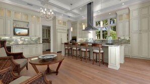 A spacious kitchen with white cabinetry, a central island with stools, and a dining area with wicker chairs. - Elevated Coastal Living | Luxury 4BR Home + Den | Designed for the Perfect Getaway (Naples)