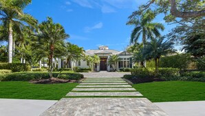 A two-story house with a paved driveway leading to the front door, surrounded by well-maintained lawns and palm trees. - Elevated Coastal Living | Luxury 4BR Home + Den | Designed for the Perfect Getaway (Naples)