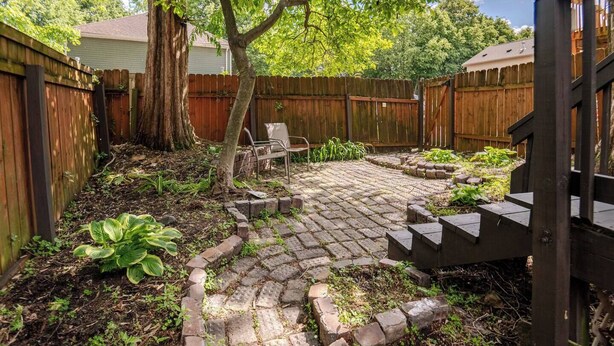 Un patio trasero con un patio de ladrillo, una valla de madera, un árbol y un banco.