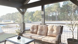 A covered outdoor seating area with a wicker sofa and a metal table, overlooking a parking lot and trees.