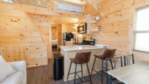 A wooden cabin interior with a kitchen, dining area, and a staircase leading to an upper level.