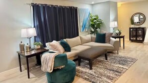 Una sala de estar con un sofá seccional color beige, un sillón verde, una mesa de centro, una alfombra, una mesita de noche con una lámpara, una planta y un espejo sobre un mueble.