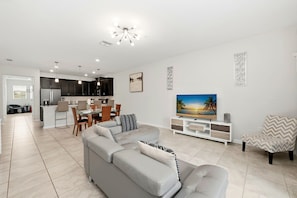 A modern living room with a grey sectional sofa, a dining area with a table and chairs, a kitchen with dark cabinets, and a TV on a white stand. - Family townhome w/ resort access (Davenport)