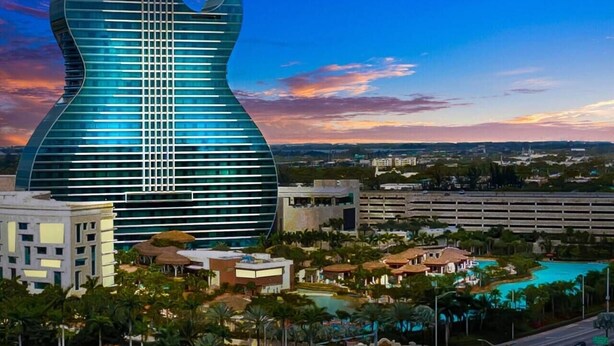 Un edificio moderno con un diseño único en forma de guitarra, rodeado de zonas ajardinadas con piscinas y vegetación.