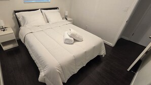 Una cama bien hecha con sábanas blancas y una toalla enrollada, una mesita de noche con una lámpara y un buró con un teléfono.