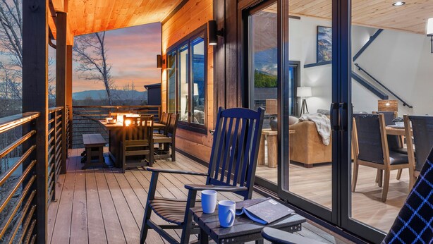 Una terraza de madera con una mesa y sillas, una silla con un libro y una taza, y vista del interior a través de puertas de cristal.
