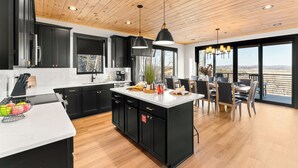 Una cocina moderna con armarios negros, encimera blanca y una isla central. Hay una zona de comedor con una mesa y sillas, y vistas al exterior a través de grandes ventanales.