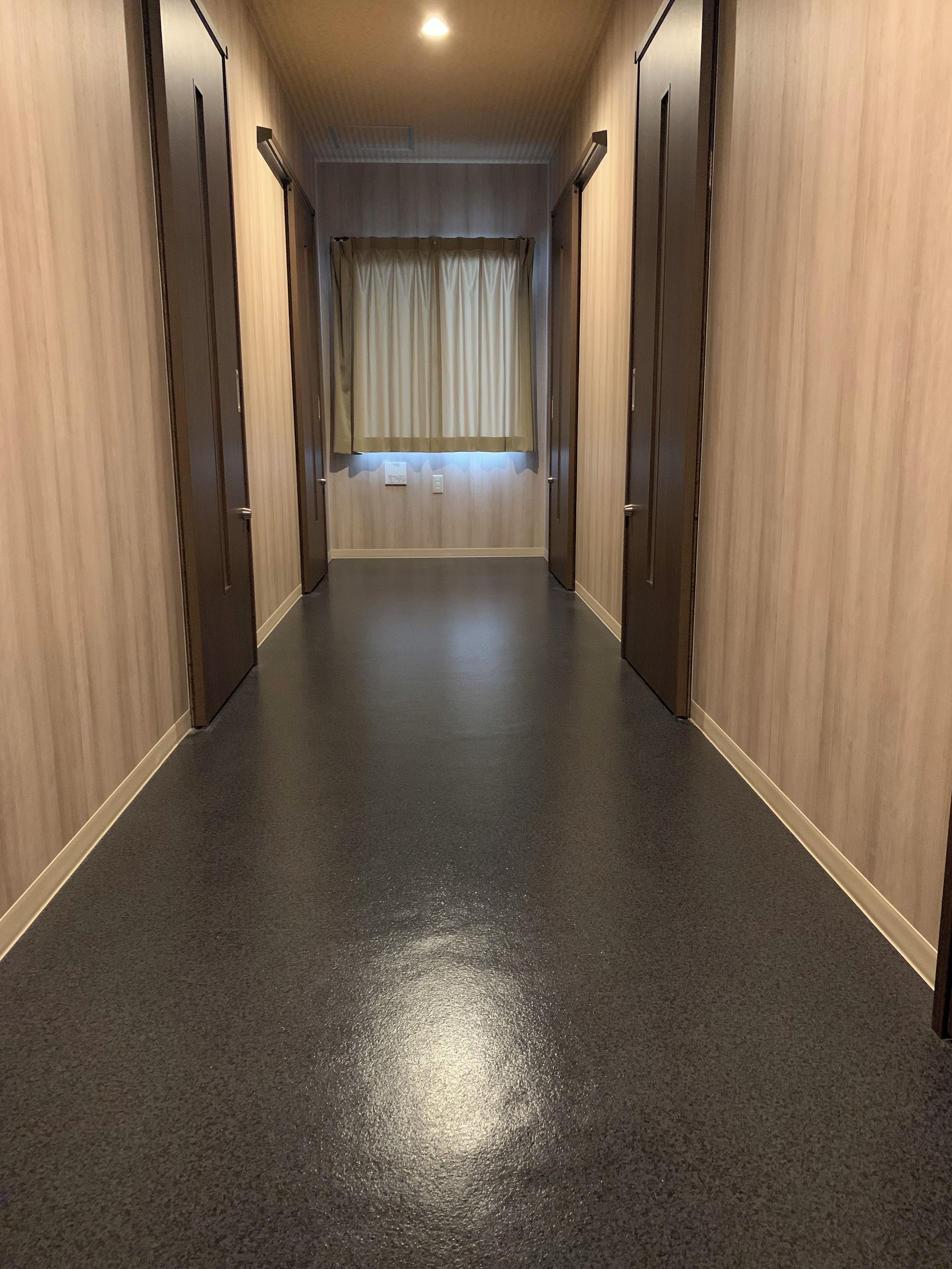 A corridor with wooden walls and a dark floor, leading to a room with a window and curtains.