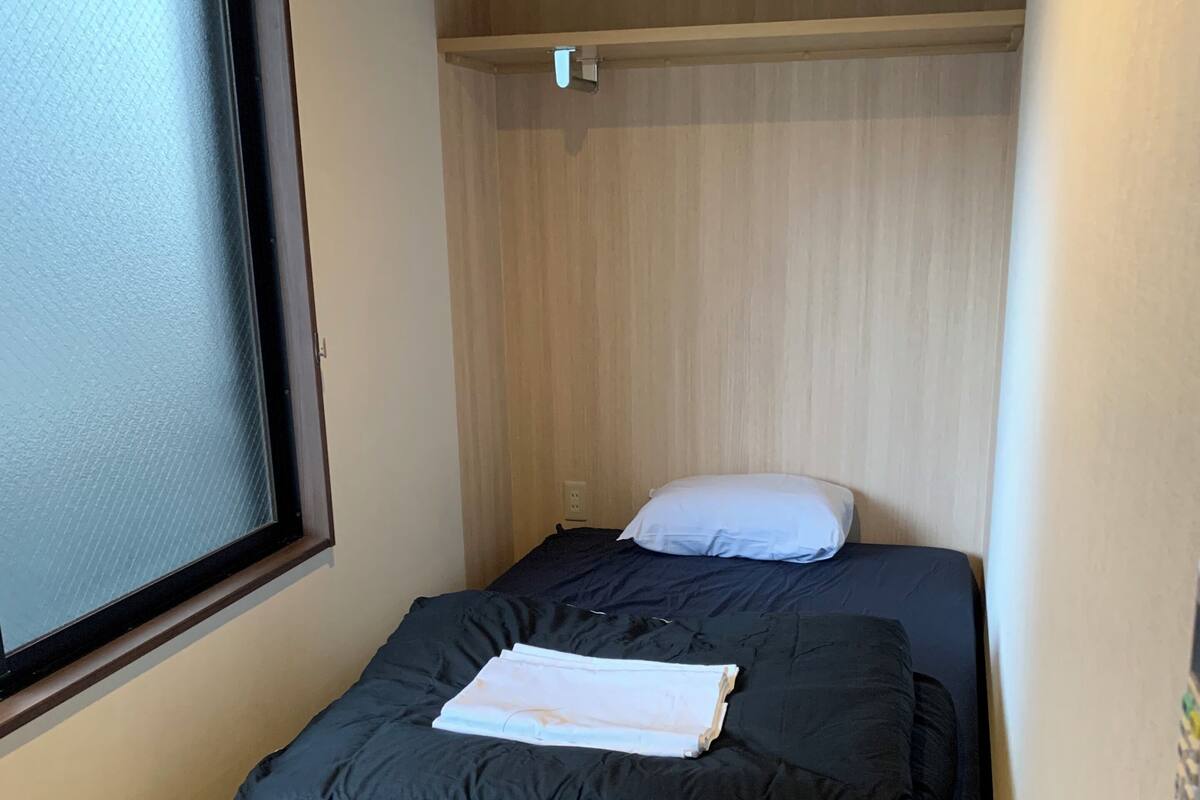 Une petite chambre avec un lit simple, une fenêtre, une tête de lit en bois et une table de chevet.