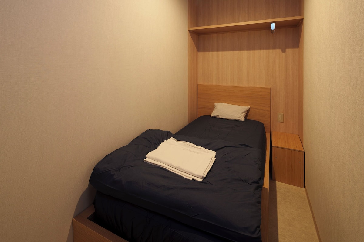 Un petit lit bien fait, avec une couverture bleu foncé et un oreiller blanc, accompagné d’une table de chevet en bois sur laquelle se trouve une petite lampe.