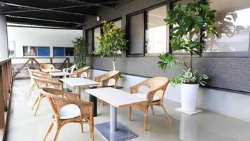A modern indoor seating area with wicker chairs and white tables, large windows, and potted plants.