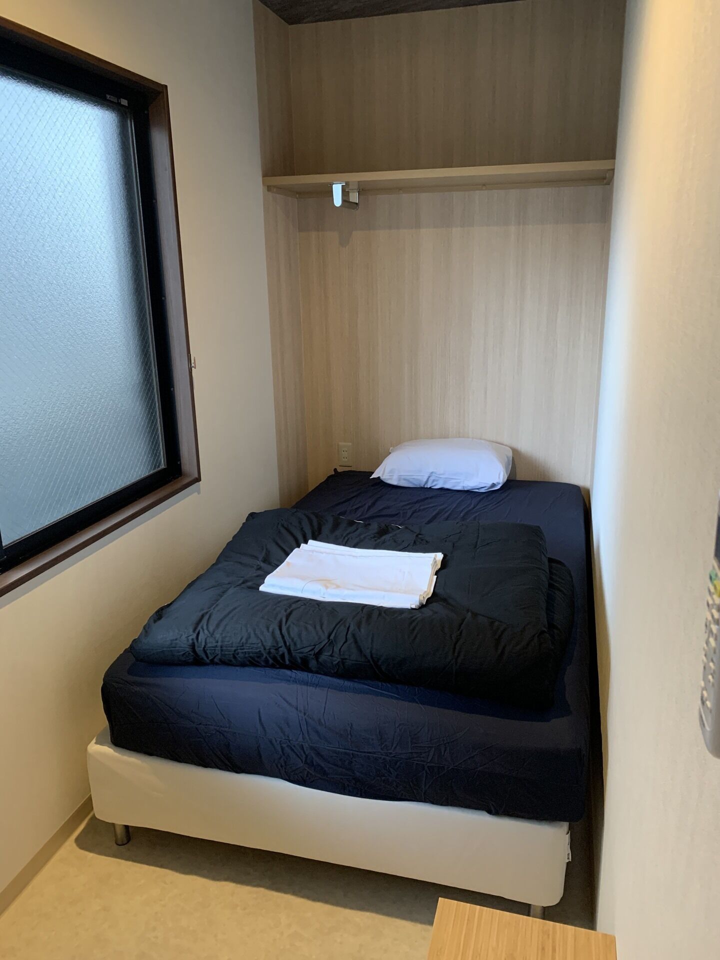 Une petite chambre avec un lit simple, une fenêtre, une étagère en bois et une prise de courant.
