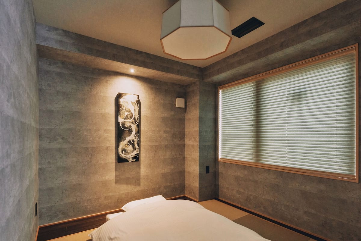 A modern bedroom with a bed, a wall-mounted artwork, a window with blinds, and a ceiling light fixture.
