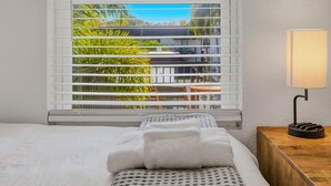 A hotel room with a bed, a window with blinds, a bedside table with a lamp, and a view of greenery outside. - Stylish Furnished 2BR in South Tampa | King Bed, Pool (Tampa)