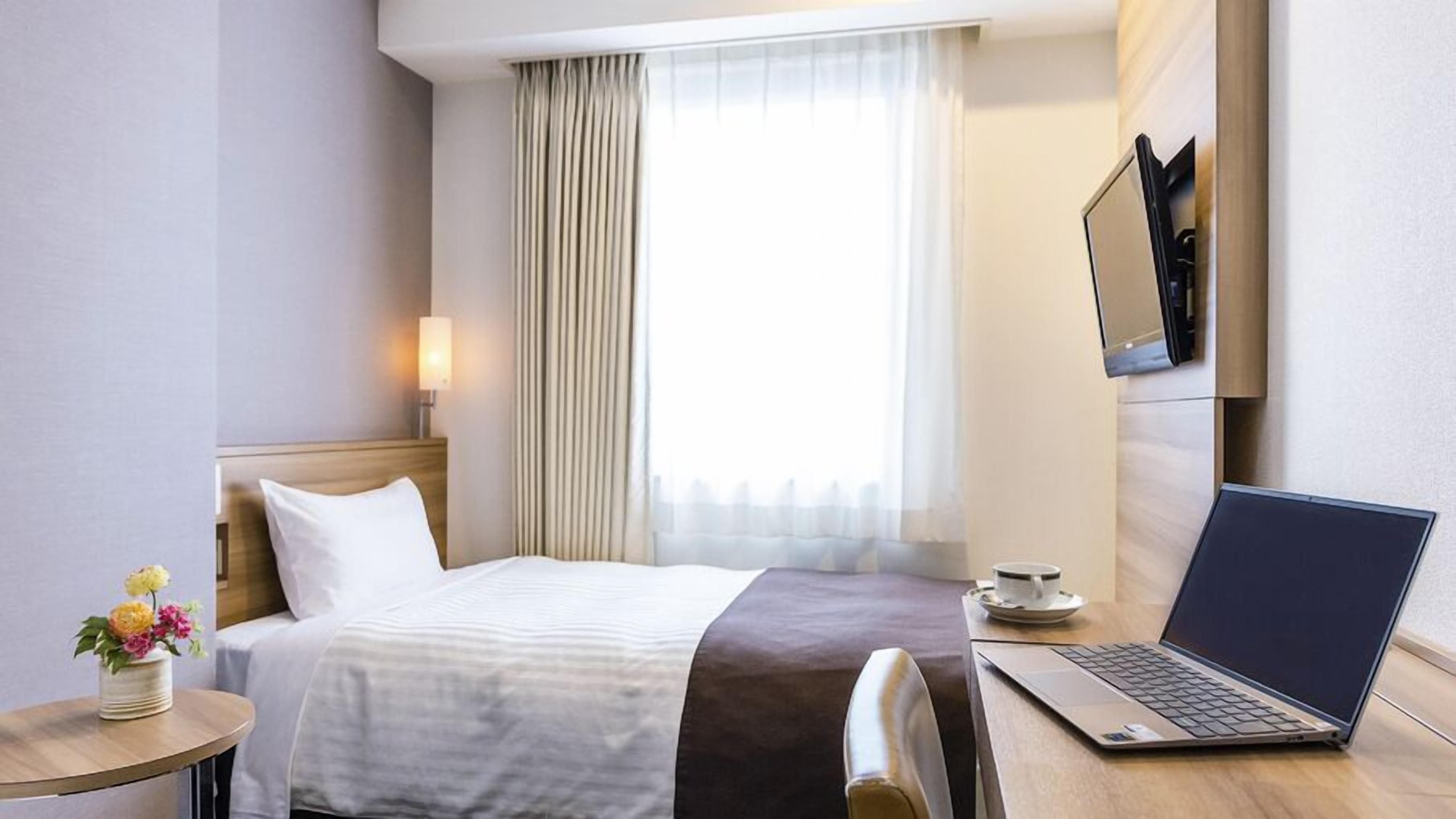 A hotel room with a bed, a laptop, a cup of coffee, and a TV on the wall.