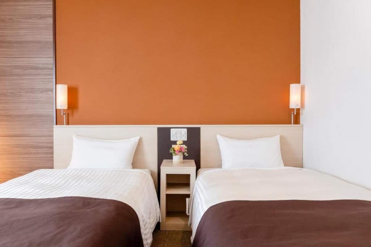 A hotel room with two beds, a nightstand with a lamp, and a wall-mounted light fixture.