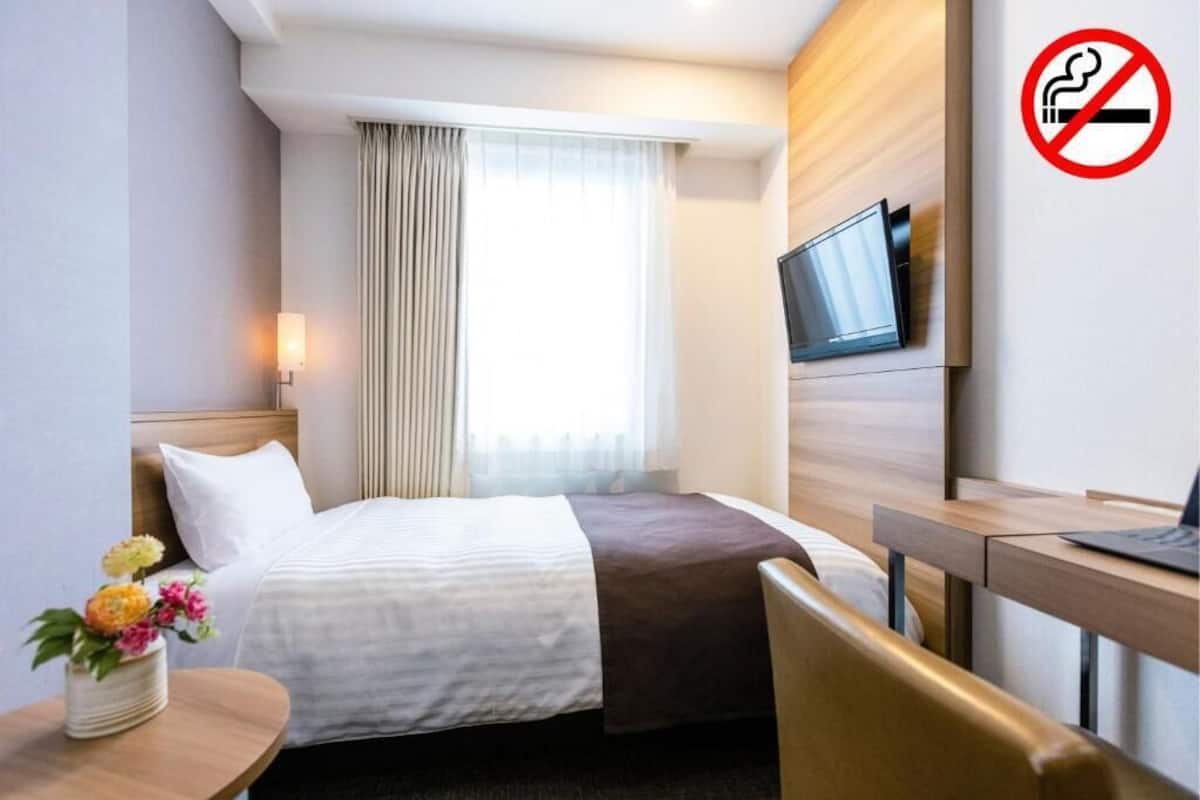 A hotel room with a bed, a desk, a TV mounted on the wall, and a no-smoking sign.