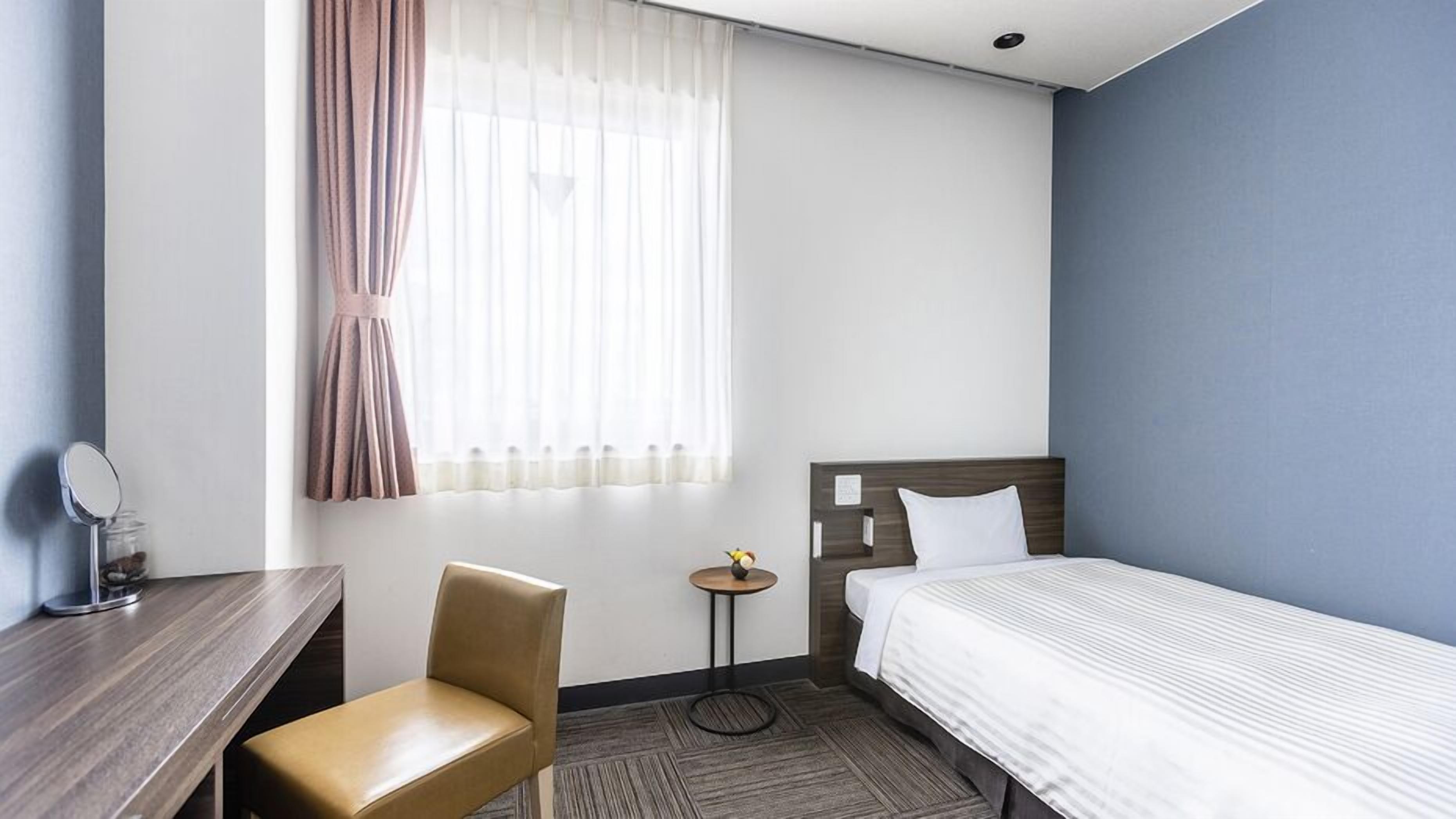 A hotel room with a bed, a chair, a small table, a mirror, and a window with curtains.