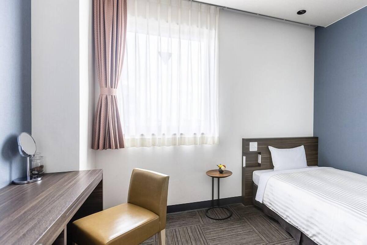 A hotel room with a bed, a chair, a small table, a mirror, and a window with curtains.