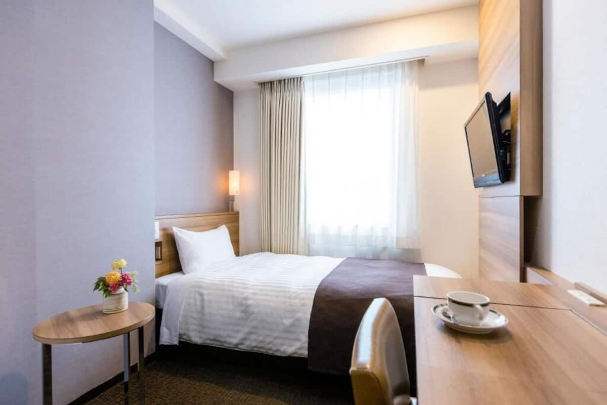 A hotel room with a bed, a bedside table with a cup, a small table with flowers, a TV mounted on the wall, and a window with curtains.